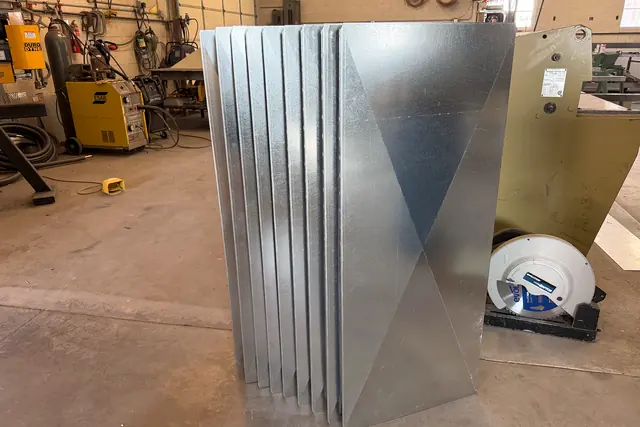 A stack of galvanized duct panels on the shop floor, each with an X-shaped cross-break pressed in.