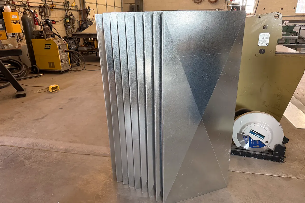 A stack of galvanized duct panels on the Macias shop floor beside the welding bay, each with an X-shaped cross-break pressed in to stiffen it.