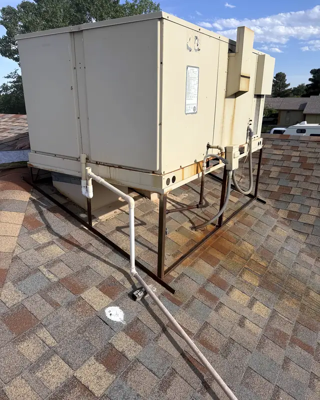 An old beige packaged rooftop unit on a rusted steel stand on a shingled house roof.
