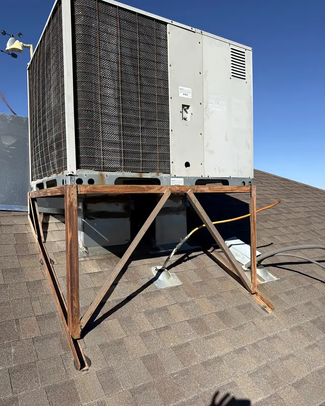 An old rooftop unit raised on a rusted angle-iron stand on a dark shingled roof, desert behind.