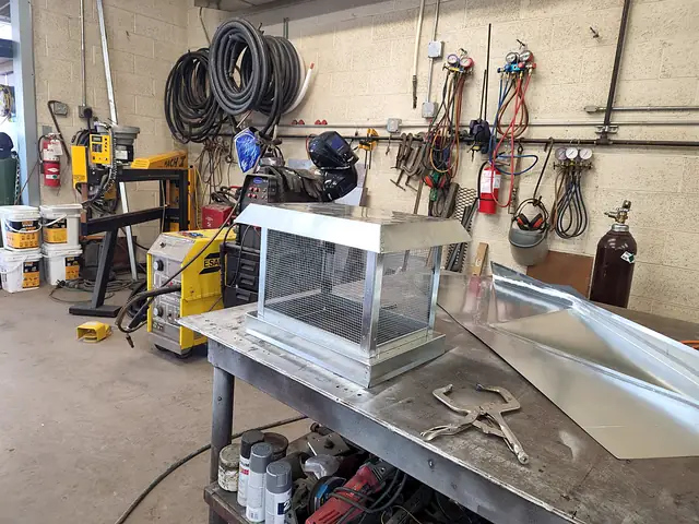 A metal chimney cap with mesh screen sides on the shop bench.