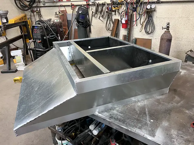 A galvanized curb adapter with two openings and a sloped side, on the shop bench.