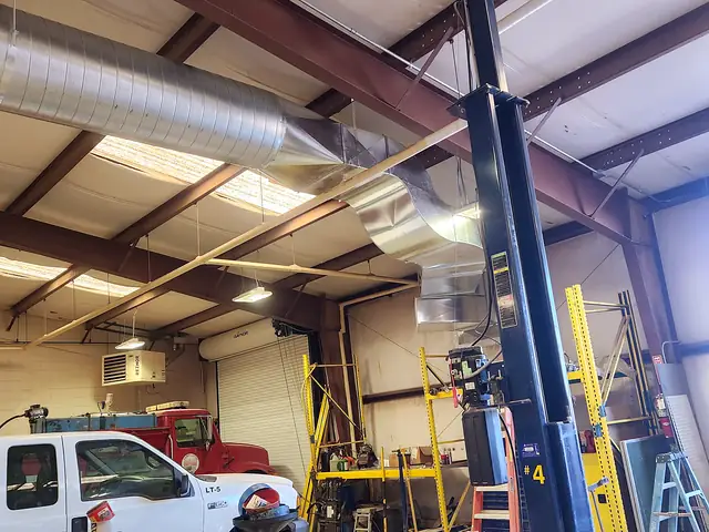 Spiral duct along the trusses of a large vehicle shop, with work trucks parked below.