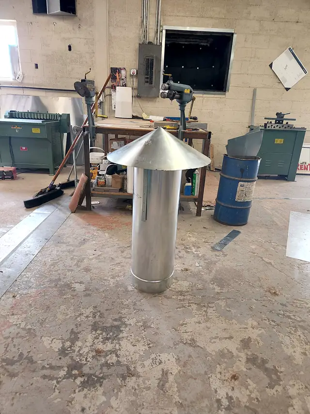 A tall round exhaust stack with a cone cap, made for a commercial hood, on the shop floor.