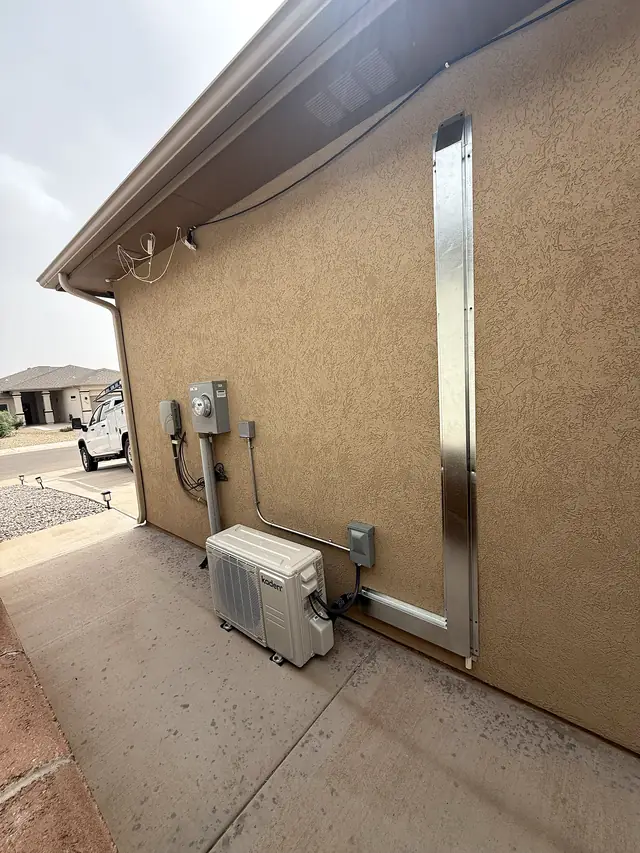 A mini-split condenser beside a stucco house, with a metal line-set cover running up the wall.