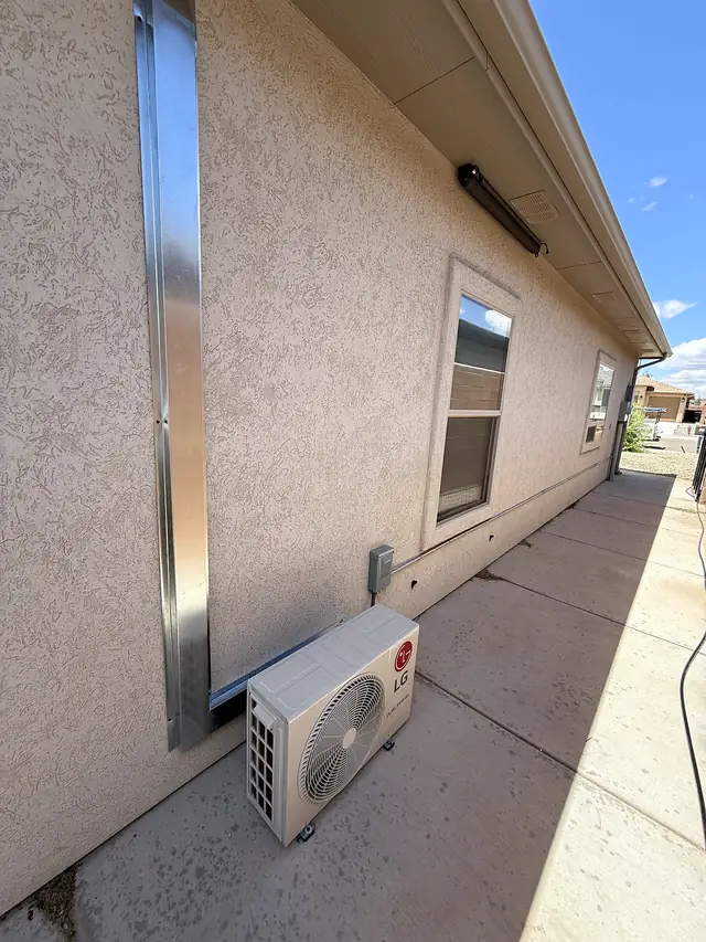 A wall-mounted mini-split condenser along a stucco house, line set covered in metal.
