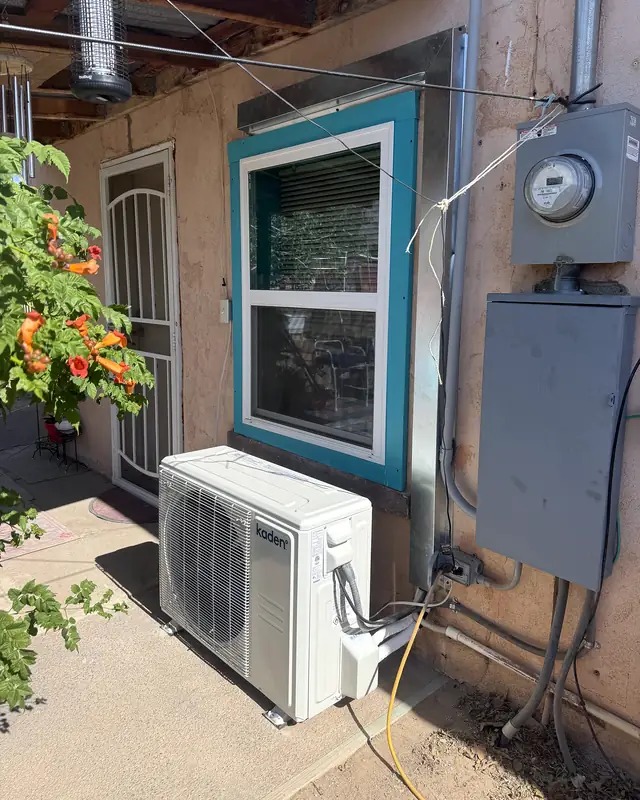 A ductless mini-split condenser on the ground beside a stucco house with a turquoise window frame.