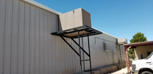 A swamp cooler on a wall-mounted steel stand on the side of a metal building.