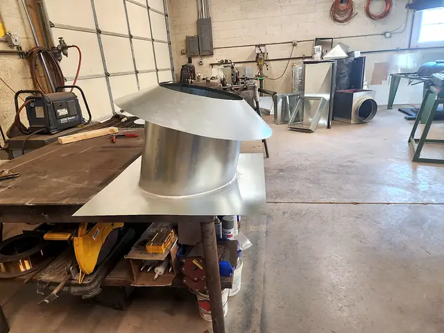 A round vent cap on its flat roof flashing, on the shop bench.