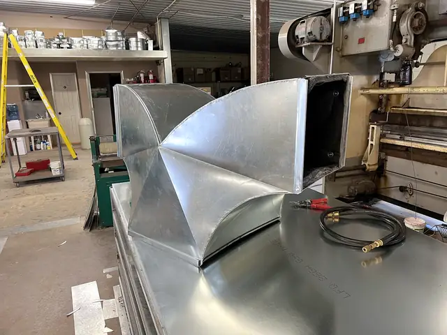 A galvanized Y fitting splitting one rectangular duct into two curved branches, on the shop table.